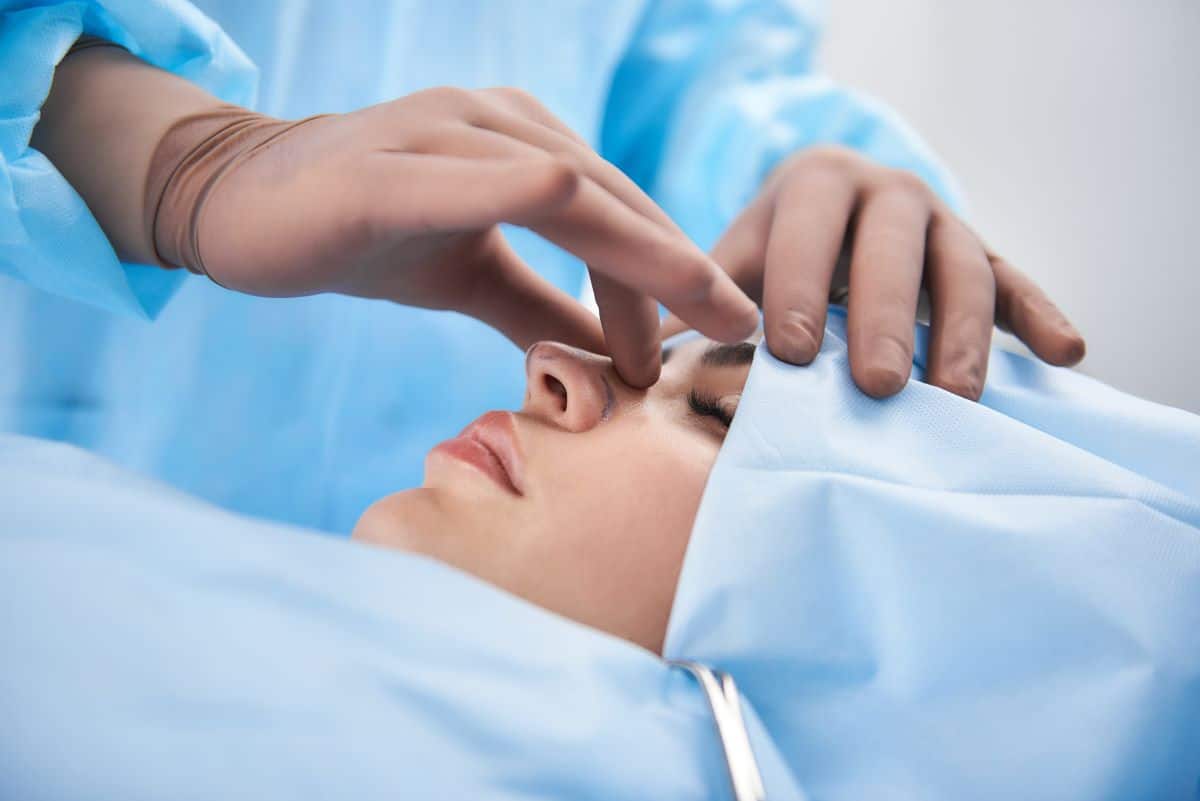 doctor's hands in rubber gloves are gently squeezing the bridge of a woman under a surgery cover with face exposed 