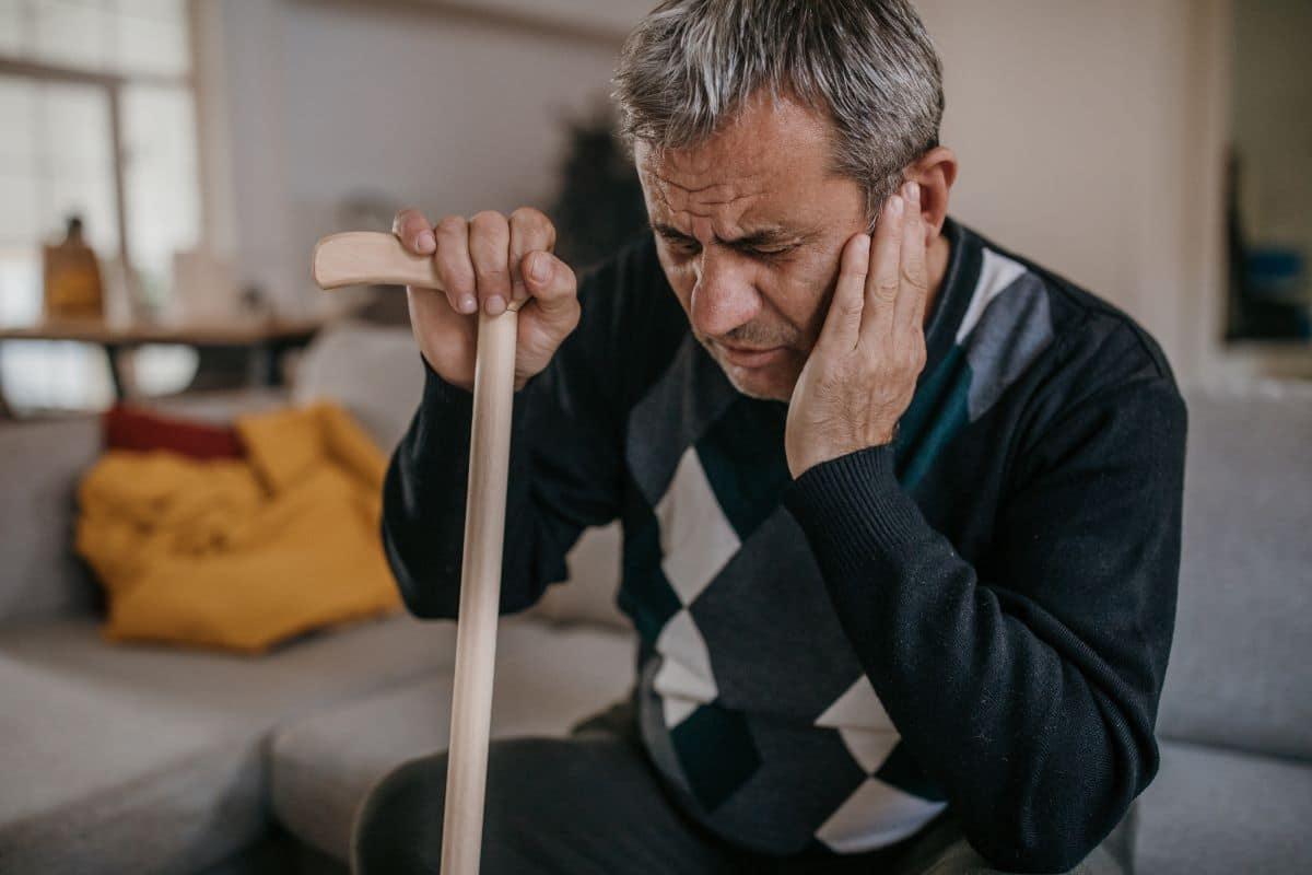 Older man sitting on the couch and holding himself upright with his cane while holding his ear due to pain from a cholesteatoma