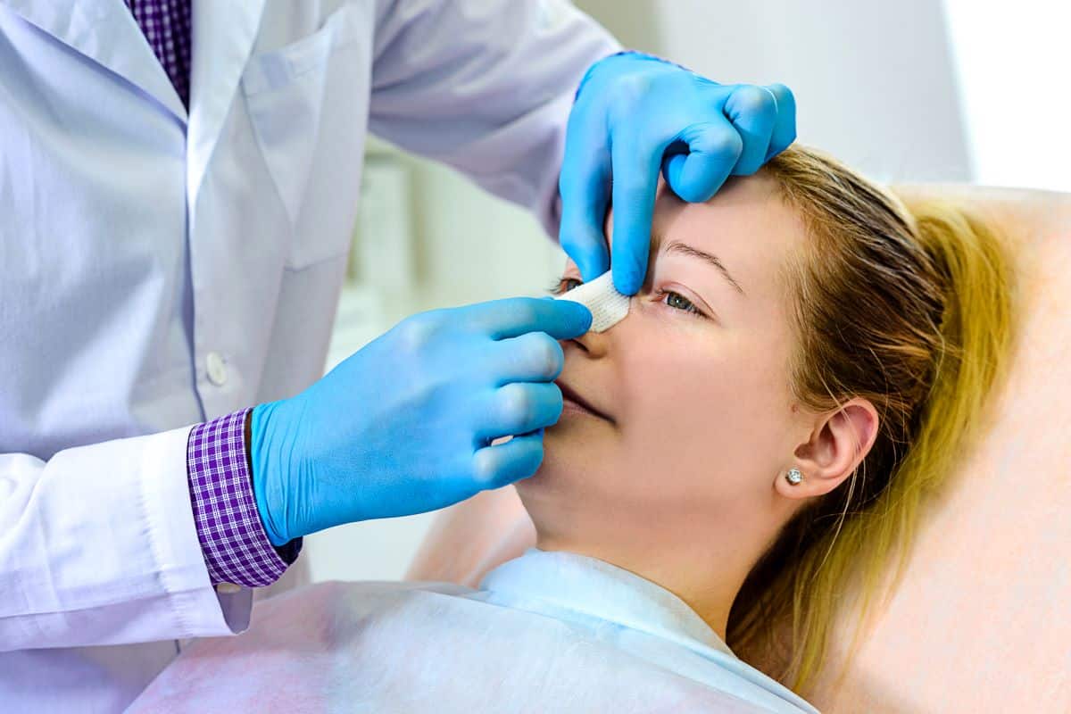 doctor wearing blue rubber gloves is placing a bandage over the bridge of a female patients nose