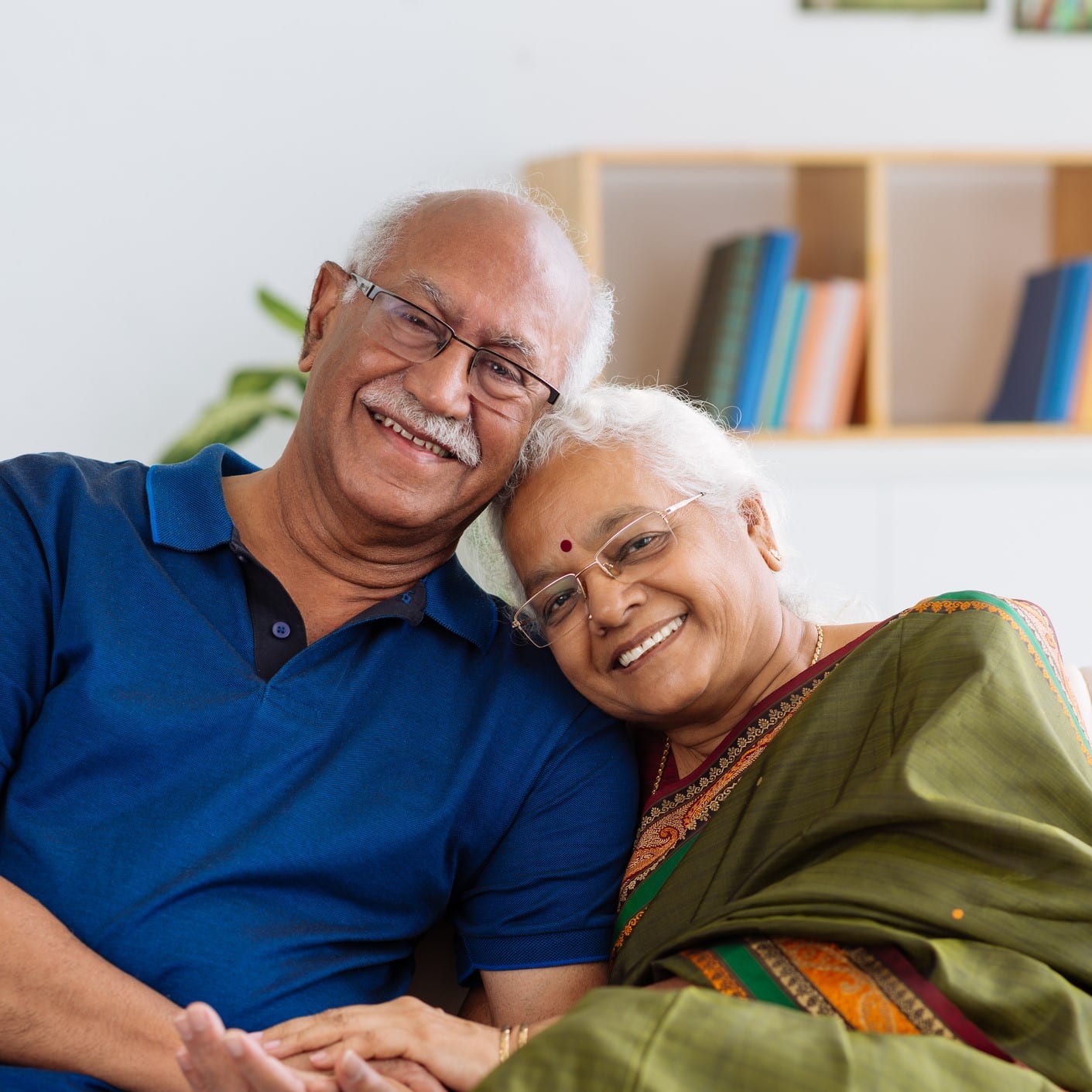 older indian man with older indian woman on a white couch. He is wearing a blue tshirt and she is wearing a green sari. They are both smiling.